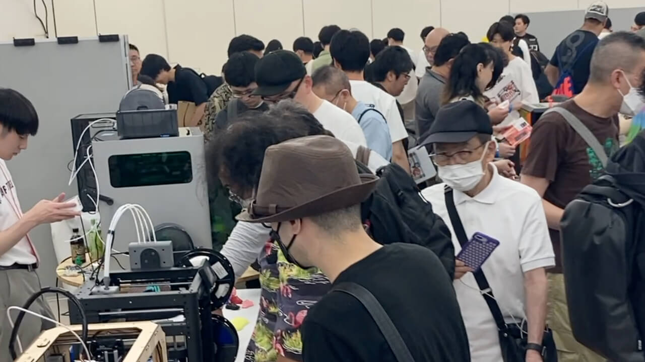 A packed exhibition floor at JRRF2025 — attendees gather around 3D printers on display.